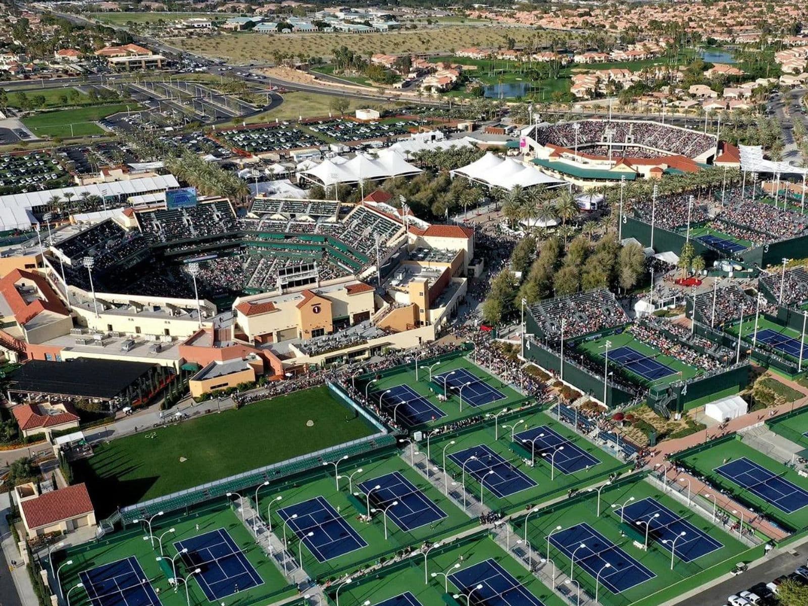 Numerous tennis courts in a natural landscape with water and trees, with a large stadium in the background.