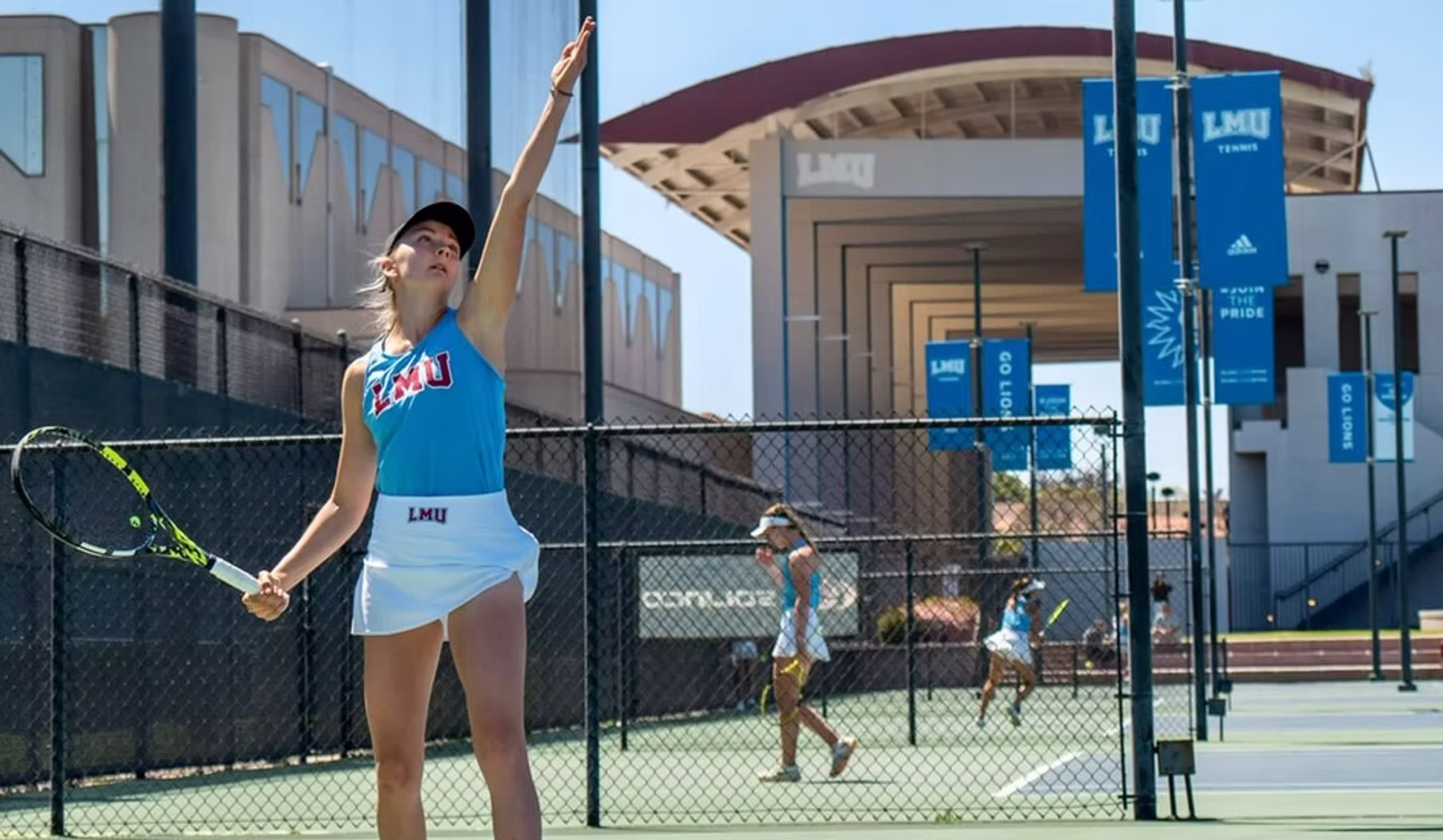 Tennisspielerin in College-Outfit beim Aufschlag auf einem Außenplatz — Banner der Universität am Zaun.