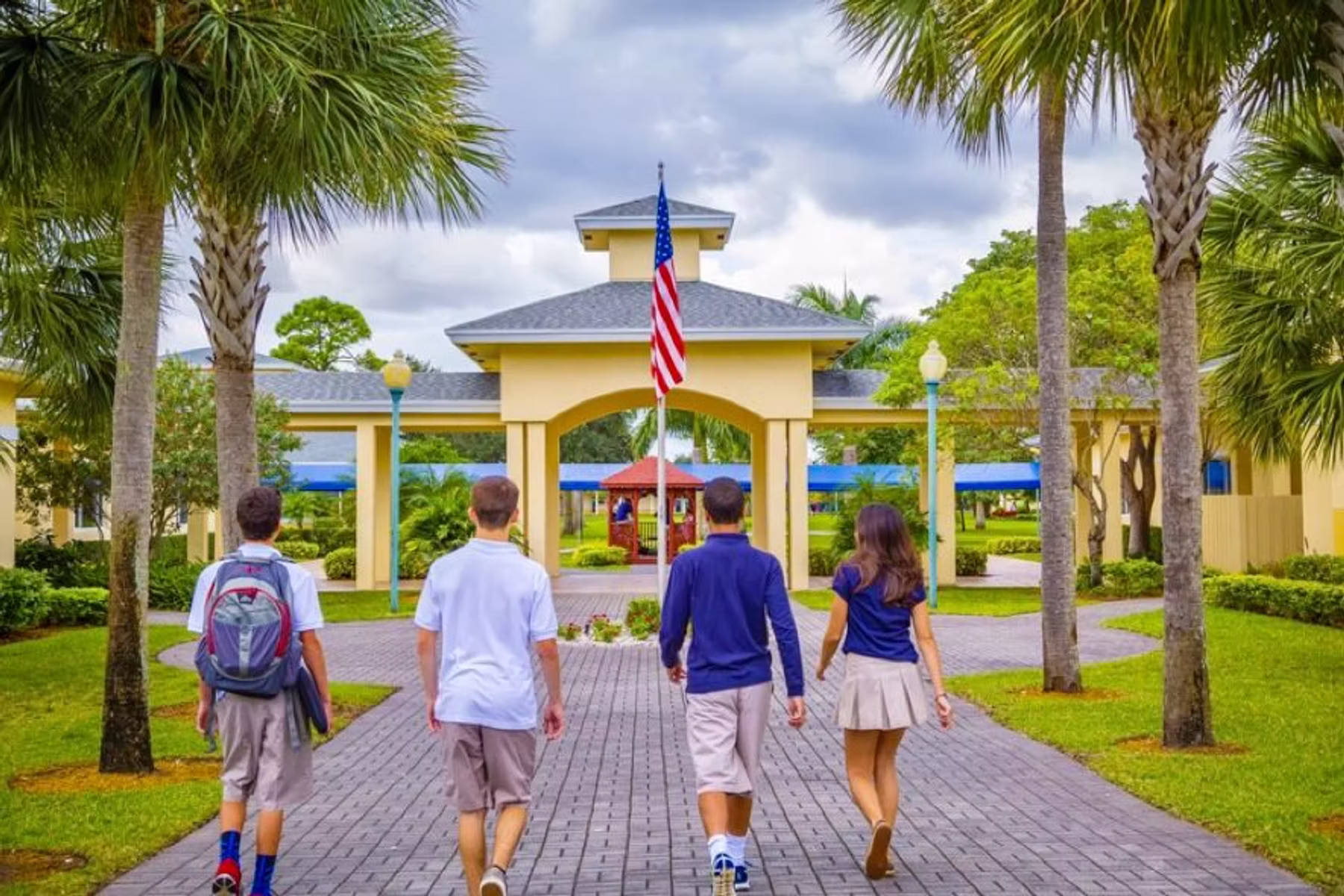 Vier Schüler auf einem gepflasterten Weg Richtung Campusgebäude mit US-Flagge, Palmen und Rasenflächen.